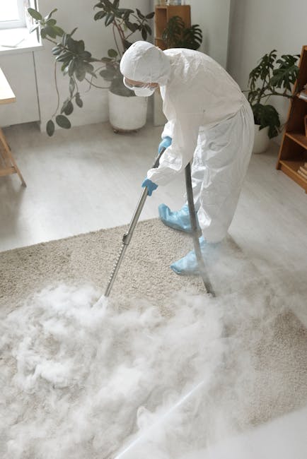 A person using a red handheld vacuum cleaner to clean a light beige textured rug on a wooden floor in a residential living room. Part of a wooden sideboard and a light-colored chair are visible in the